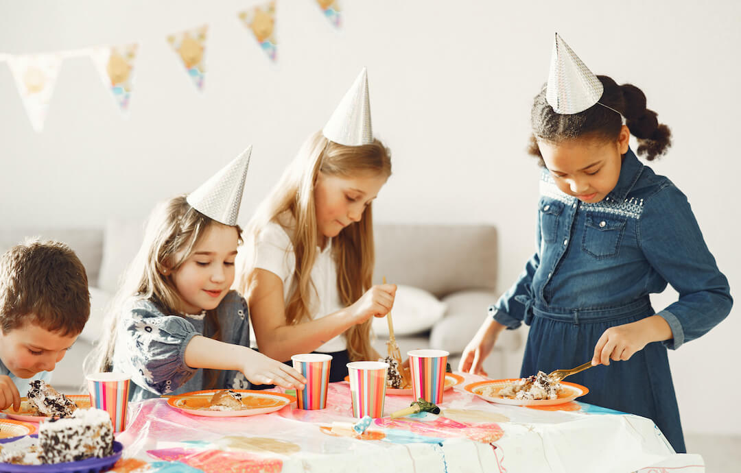 foto de niños y niñas en una fiesta de cumpleaños. llevan gorros, hay banderines y están en torno a una mesa con tartas y comidas sonriendo