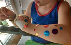 mi hijo con 4 años sentado frente a una mesa despegando gomets de colores de su brazo izquierdo que tiene el puño vendado