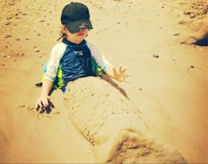 mi hijo sentado en la playa, con gorra y camiseta protectora, y con las piernas cubiertas de arena en forma de cola de sirena