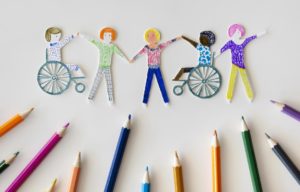 dibujo de varias personas dandose las manos, algunas en silla de ruedas. Debajo están los lápices de colores con los que se hicieron.