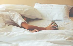 mujer durmiendo, con la cabeza debajo de la almohada y cogiendo el despertador con una mano