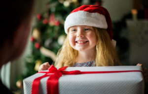 niña rubia sonriente con un gorro de papa noel, delante de un arbol de navidad, entrega un regalo envuelto con un lazo rojo a un adulto