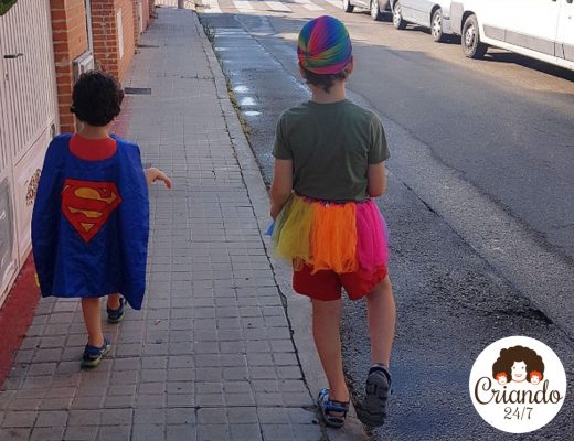 mis hijos de espaldas, caminando por la acera, uno lleva una capa de superman y el otro va con un tutú y una gorra multicolor
