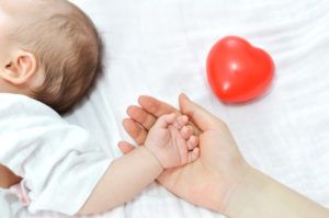 mano de una madre que sostiene la de un bebé recostado sobre una cama de sábanas blancas y un corazón de goma rojo