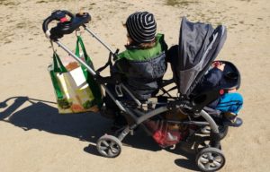mis hijos con 2 y 4 años en un carro gemelar. van sentados espalda con espalda, se ve una bolsa de mercadona colgada, el pequeño tiene la capota puesta