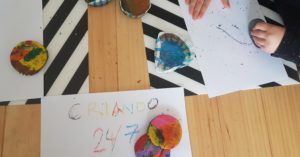 mi hijo de 6 años pintando sobre un folio blanco con una cera reciclada con forma circular multicolor. se ve un folio donde dice criando 24/7