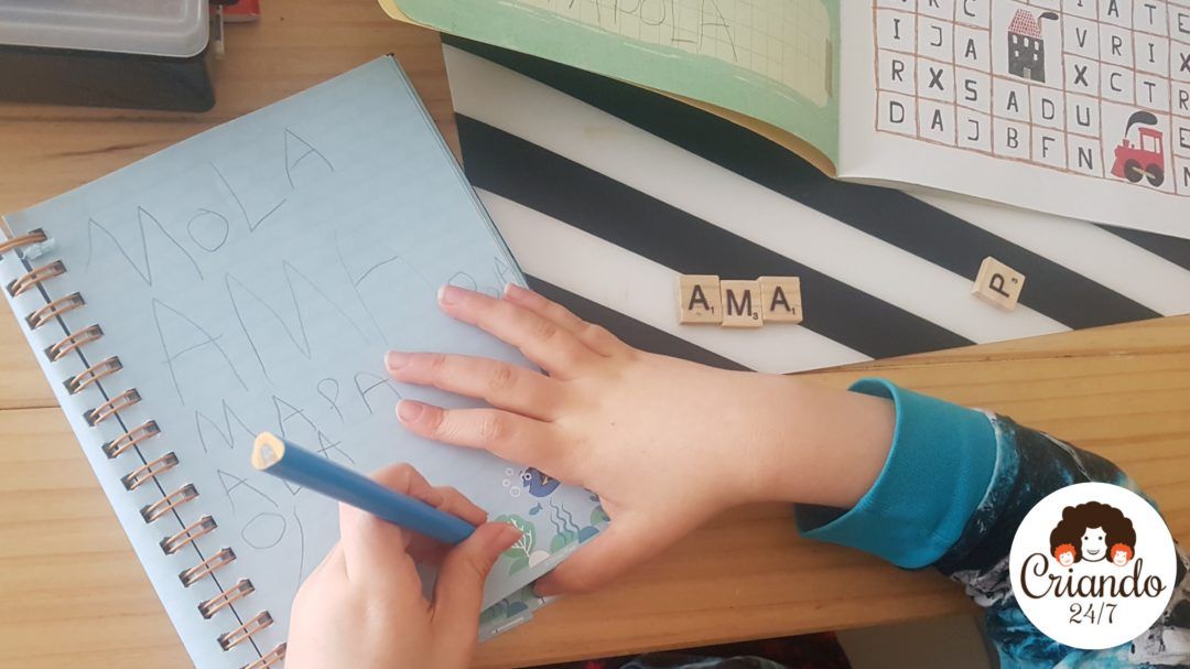 mi hijo de 6 años escribiendo en un cuaderno con un lapiz, a su lado unas letras de scrabble de ,adera con la palabra AMA. logo de criando 24/7