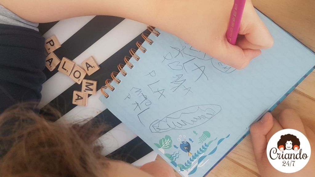 mi hijo de 8 años con hemiparesia derecha, escribiendo con la mano izquierda en su cuaderno usando un lapiz gordo con forma triangular, y varias letras de madera de scrabble a su lado. logo de criando 24/7
