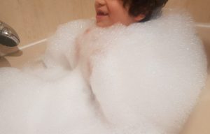 mi hijo de 6 años rodeado de espuma blanca hasta el cuello, mientras sonríe en la bañera