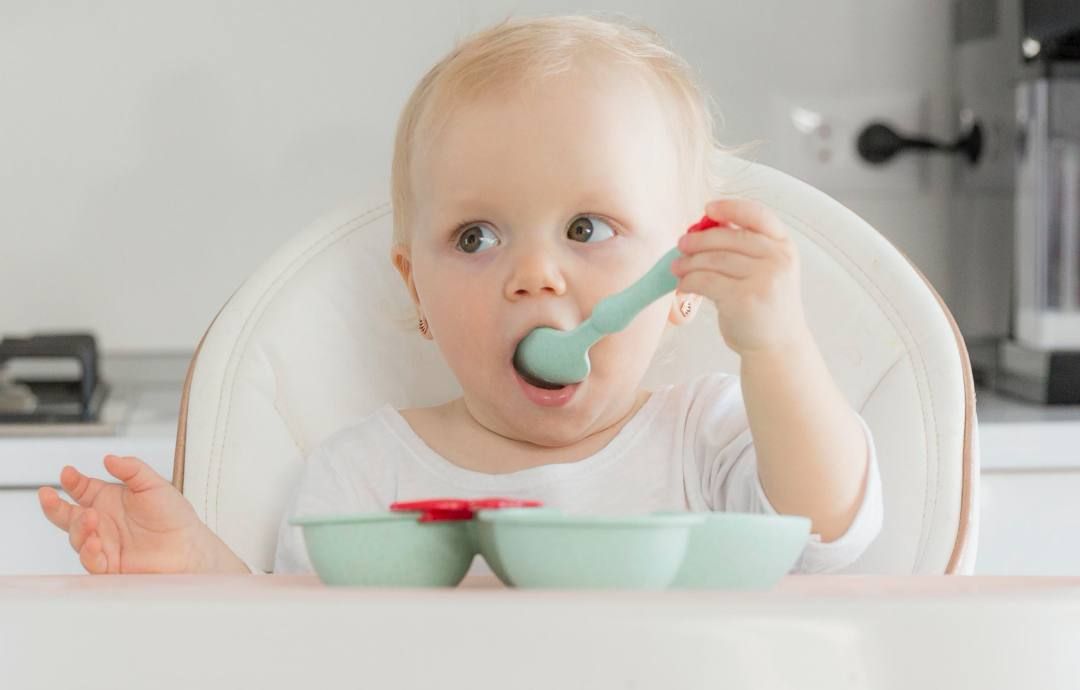 bebé rubio sentado en una trona comiendo con una cuchara