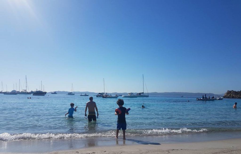 mis hijos de espaldas en la orilla del mar de Cerdeña con unos barcos de fondo