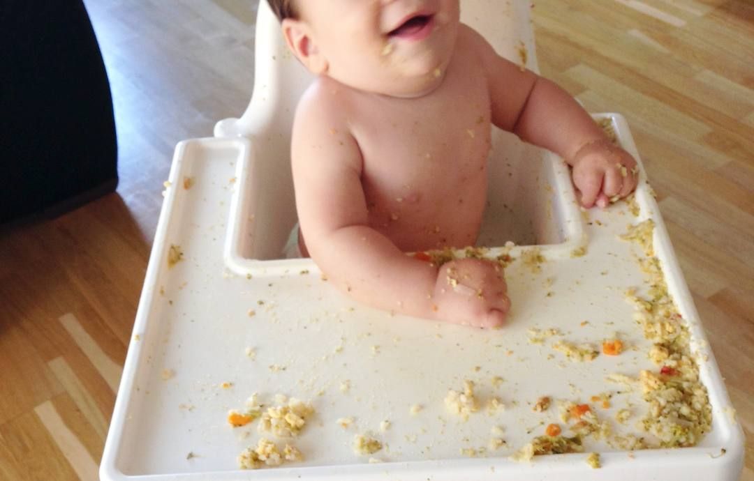 mi hijo pequeño, de bebé, sentado en la trona mientras sonríe. la bandeja está llena de comida desparramada.