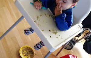 mi hijo pequeño comiendo con las manos sentado en la trona, en el suelo se ve el plato que ha lanzado