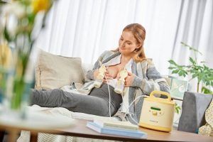 mujer sentada en sofá mientras se extrae leche con un sacaleches eléctrico doble que sujeta con las manos en cada pecho
