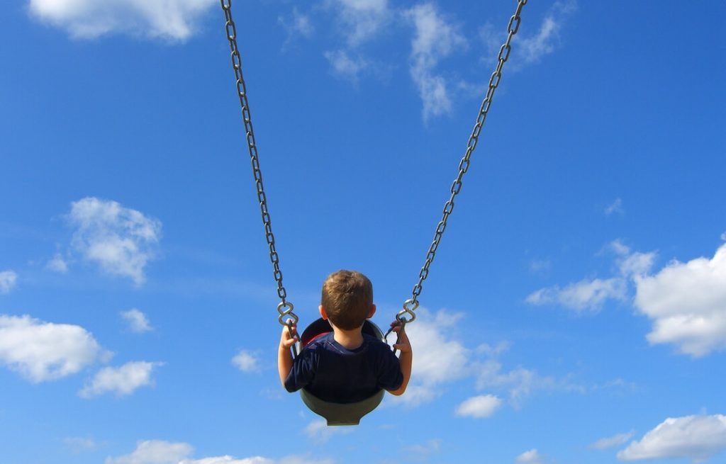 foto de un niño de espaldas columpiándose con el cielo azul con nubes blancas de fondo