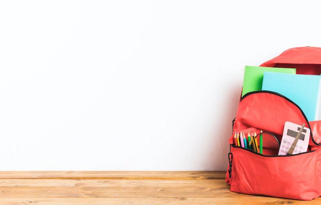 Mochila roja abierta con material escolar apoyada en un suelo de tarima.