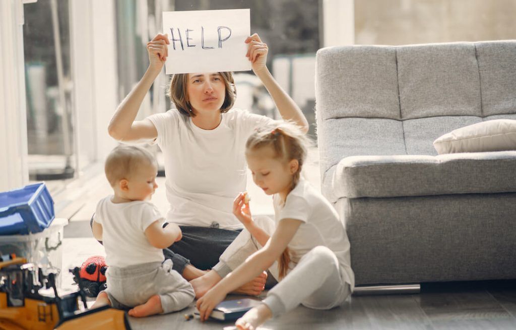 madre con 2 niños pequeños sentados en el suelo del salon. la madre tiene cara de agotada y sostiene en alto un cartel que dice HELP