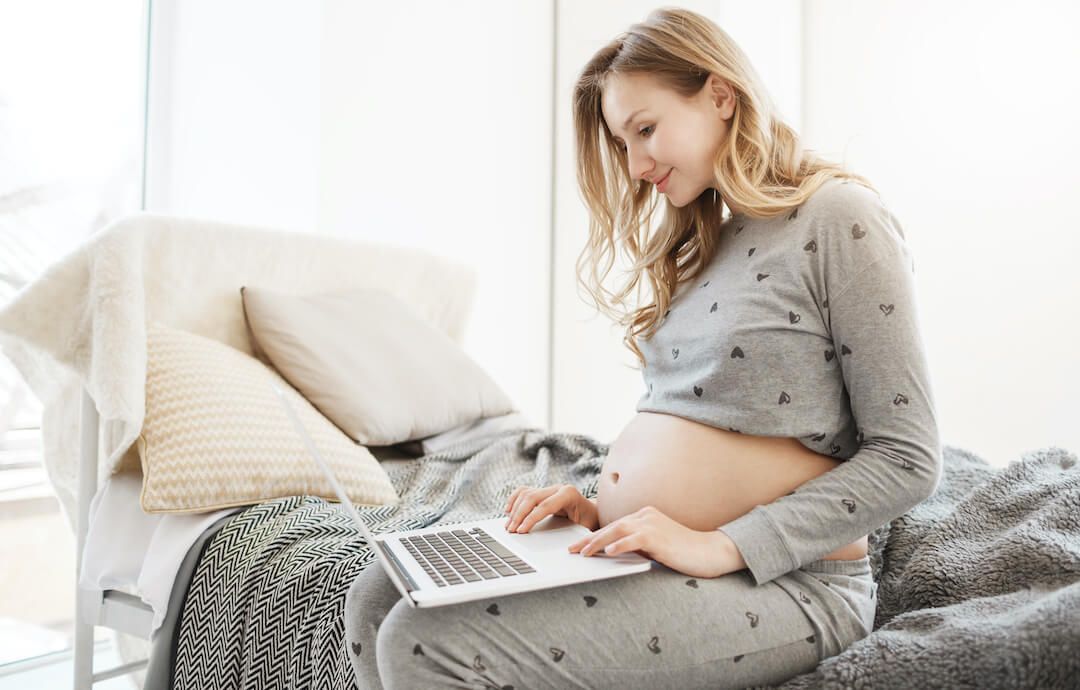 mujer embarazada sentada en el sofa mirando la pantalla de su portatil mientras sonríe