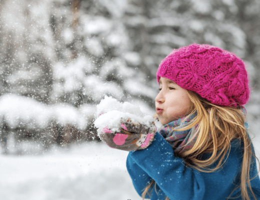 niña rubia con gorro de lana y abrigo, solplando hacia un montón de nieve que tiene en sus manos. De fondo se ve un bosque nevado.
