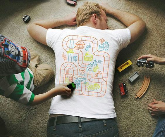 un padre recostado en el suelo boca abajo, con una camiseta que tiene dibujado un circuito para coches. Se ven unos unos niños con coches de juguete jugando sobre este circuito.