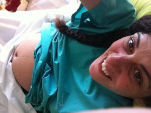 selfie recostada en una cama de hospital, con bata verde desde la que asoma mi tripa de 33 semanas, mientras sonrío