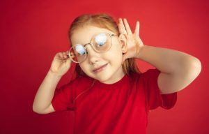 niña pelirroja con gafas con la mano detrás de la oreja haciendo un gesto de oir