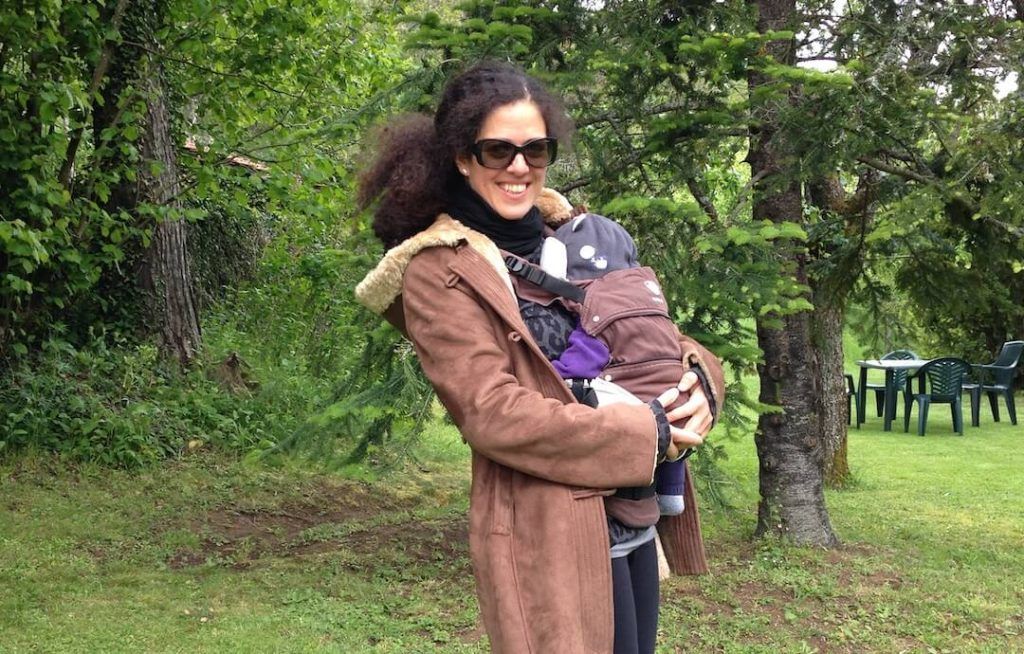 foto en la que se me ve abrigada en un jardín, porteando mi hijo pequeño en una mochila ergonómica Manduca