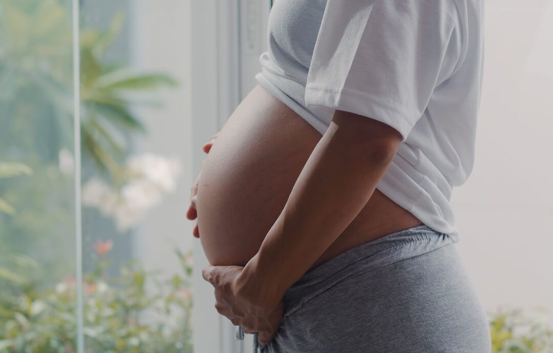 mujer embarazada cogiéndose la tripa frente a un ventanal donde se ven plantas.