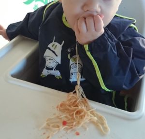 mi hijo sentado en una trona de ikea, con un babi con mangas, comiendo espaguetis con la mano