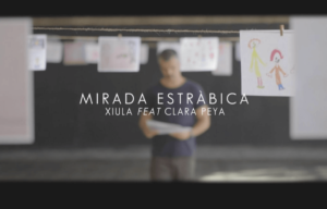 fotograma del videoclip de Xiula, donde dice MIRADA ESTRABICA y se ve al cantando con un dibujo en la mano y detrás varias cuerdas donde cuelgan dibujos en folios blancos