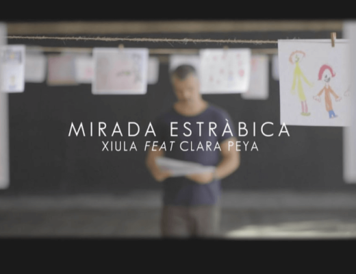 fotograma del videoclip de Xiula, donde dice MIRADA ESTRABICA y se ve al cantando con un dibujo en la mano y detrás varias cuerdas donde cuelgan dibujos en folios blancos
