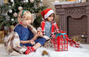 una niña con gorro de papa noel y un niño con orejeras de reno sentados delante de un arbol de navidad frente a unos regalos