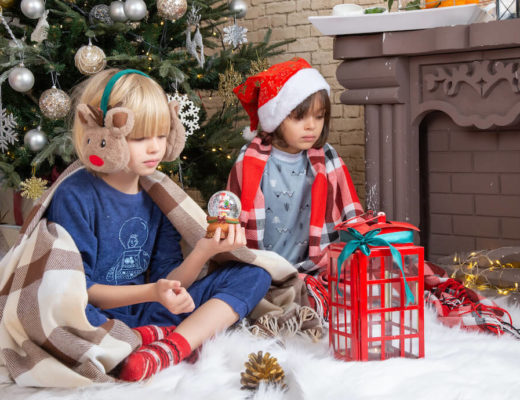 una niña con gorro de papa noel y un niño con orejeras de reno sentados delante de un arbol de navidad frente a unos regalos