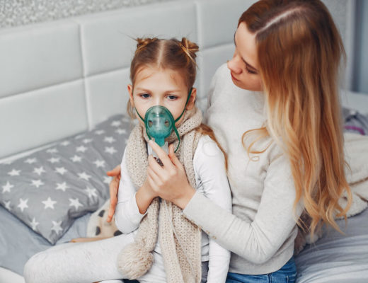mujer joven sentada en la cama junto a una niña le sostiene una mascarilla de oxigeno