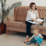 mujer joven rubia sentada en el sofa mientras habla por el movil y escribe en un portatil en el suelo a su lado hay un bebé rubio jugando