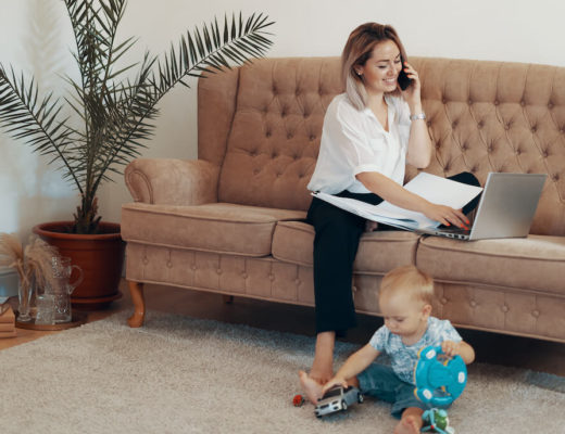 mujer joven rubia sentada en el sofa mientras habla por el movil y escribe en un portatil en el suelo a su lado hay un bebé rubio jugando