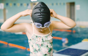 niña colocandose gafas de natación y gorra, detrás se ve una piscina olímpica
