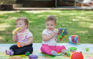 dos bebes sentados en una manta en un parque, rodeados de juguetes
