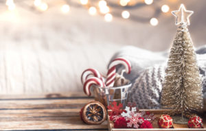 decoración navideña: un arbolito pequeño, al lado de un vaso de cristal con bastones de caramelo rojos y blancos. Detrás se ve una manta mullida color crema y guirnaldas de luces cálidas