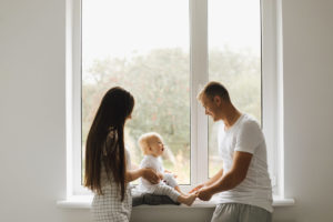 Pareja joven de hombre y mujer con un bebé sentado sobre un cojin en el murete de una ventana