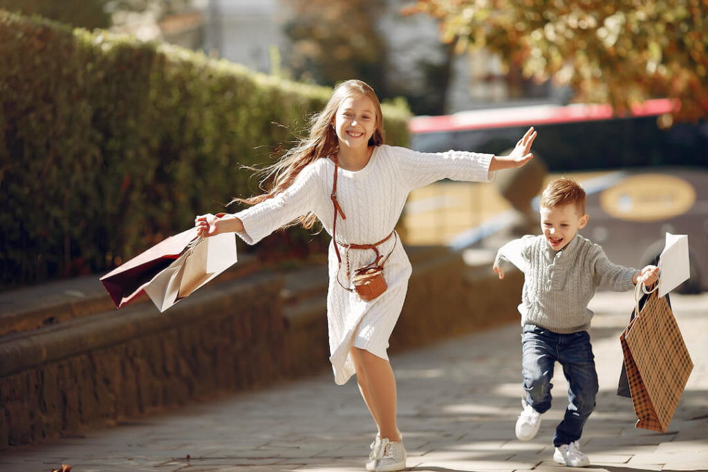 una niña y un niño corriendo por la acera, llevan bolsas de papel en sus manos