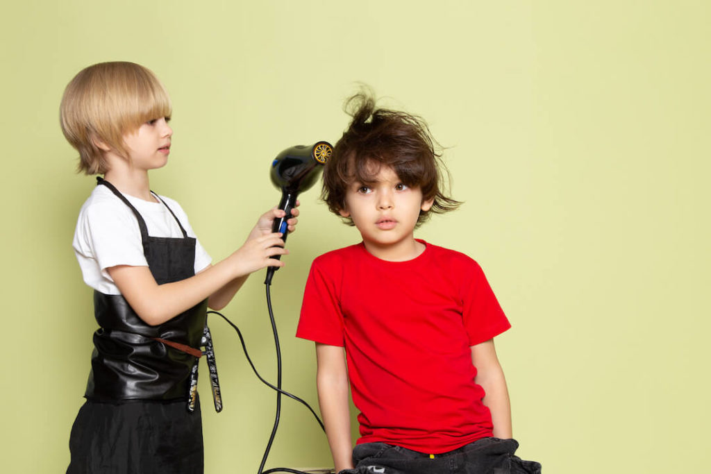 niño rubio con delantal de peluquero secando el pelo a otro niño moreno sentado