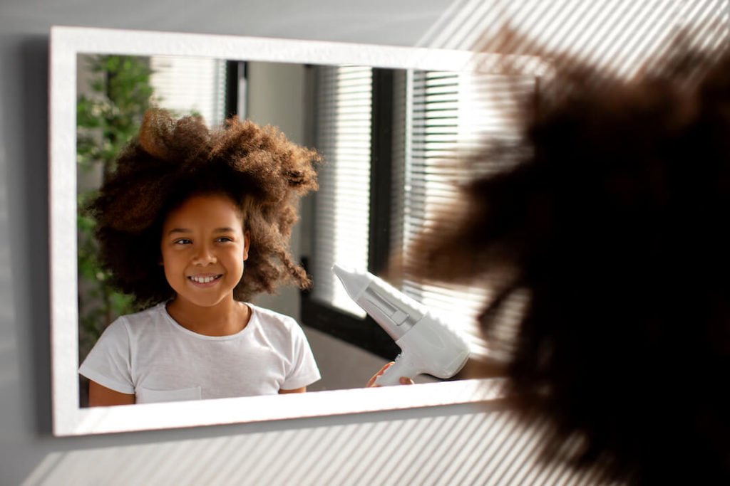 niña con pelo afro secándose el pelo frente al espejo