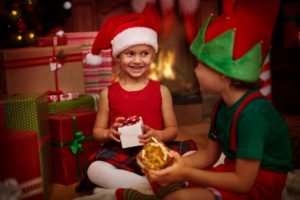 niño y niña con gorros navideños, sosteniendo regalos junto al arbol y sonriendo