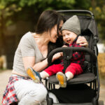 foto de una madre en cuclillas abrazando a su bebés que va en un carro, están en un parque