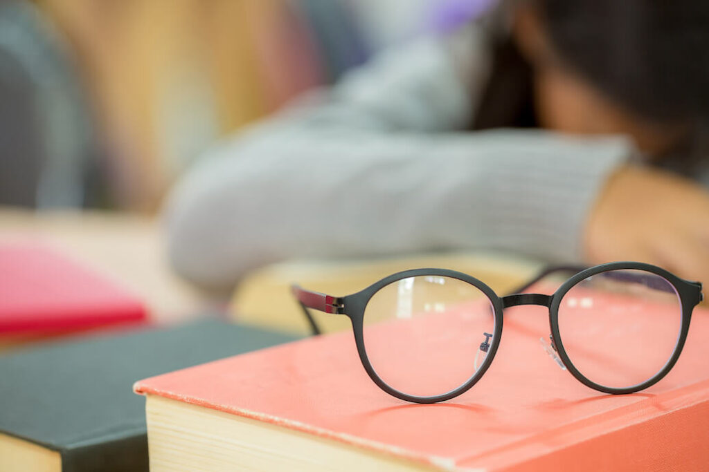 gafas sobre un libro, detrás se ve una niña recostada sobre el escritorio del colegio