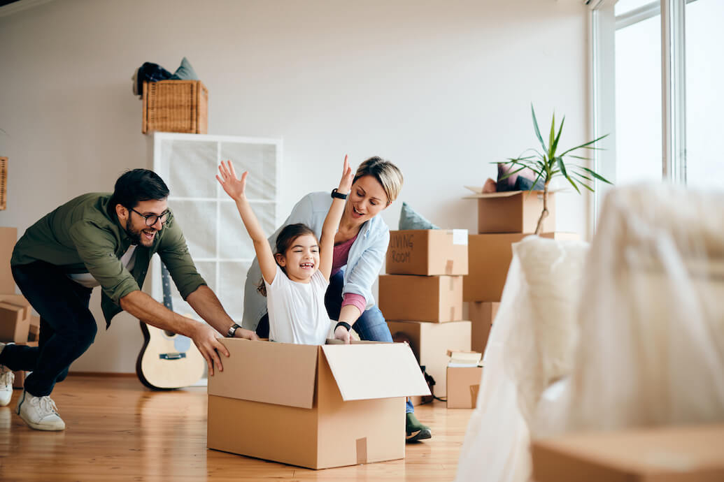 foto de una familia sonriendo con cajas de mudanza, la madre y el padre empujan a una niña que está sentada dentro de una caja con los brazos en alto