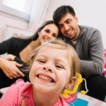 niña pequeña sonríe a cámara mostrando los dientes, detrás se ve una pareja de hombre y mujer