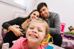 niña pequeña sonríe a cámara mostrando los dientes, detrás se ve una pareja de hombre y mujer