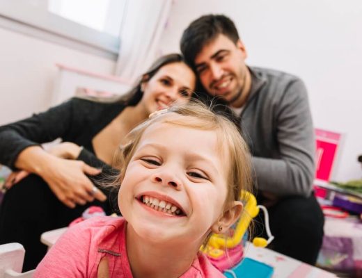 niña pequeña sonríe a cámara mostrando los dientes, detrás se ve una pareja de hombre y mujer